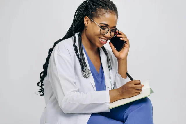Smiling healthcare professional in a white coat and scrubs taking notes for Accurate Data Entry while talking on a smartphone.