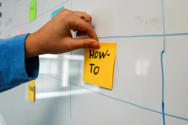 A hand placing a yellow sticky note labeled How-To on a whiteboard to organize Patient Satisfaction in Healthcare strategies.