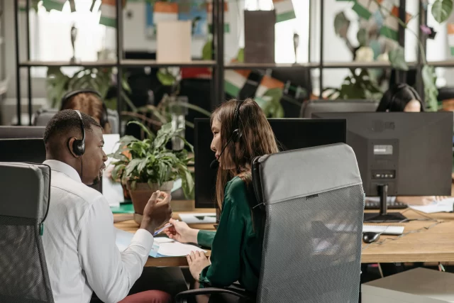 Two colleagues wearing headsets collaborate at their desk during a busy MTM pharmacy workflow.