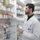 Pharmacist in a white coat selecting medication from pharmacy shelves stocked with Azimex, Amoxim, and ADCEF for Advanced Medical Supply.