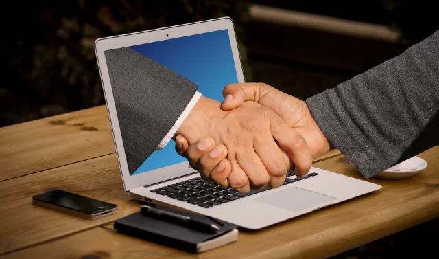 A digital handshake emerging from a laptop screen, symbolizing the remote partnership model of Switzerland's HIM.